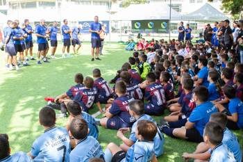 A crowd of children dressed in football uniforms, sitting on the pitch and listening to the team of coaches talking to them